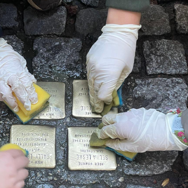 Vier Hände mit Schutzhandschuhen reinigen Stolpersteine; auf einem von ihnen ist der Name Selma Leske lesbar
